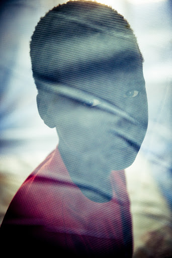 Striking portrait of a young boy with brown skin, wearing a red shirt, partially obscured by a patterned overlay creating a textured effect.