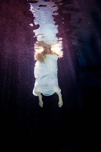 Surreal underwater view of a woman in a flowing white dress, back to the viewer, with sunlight streaming from the water's surface.