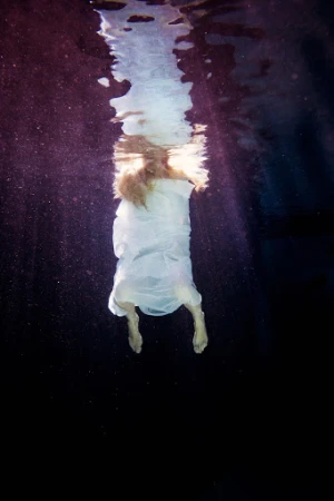 Surreal underwater view of a woman in a flowing white dress, back to the viewer, with sunlight streaming from the water's surface.