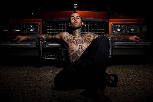 Shirtless man with tattoos poses in front of a vintage orange car with a silver grill in a dark indoor environment with a shiny floor.