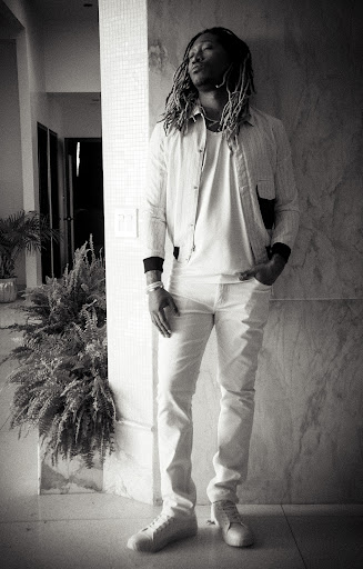 Monochrome portrait of Future with dreadlocks leaning against a marbled wall, wearing a light jacket, t-shirt, and pants.