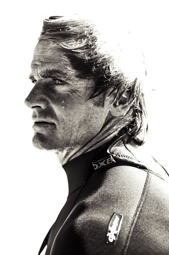 Profile view of adult male surfer with wet hair wearing a black wetsuit against a bright white background in a monochromatic photograph.