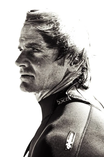 Profile view of adult male surfer with wet hair wearing a black wetsuit against a bright white background in a monochromatic photograph.