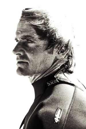Profile view of adult male surfer with wet hair wearing a black wetsuit against a bright white background in a monochromatic photograph.