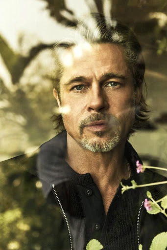 Portrait of actor Brad Pitt looking through a window; he wears a black zip-up jacket and collared shirt with a neutral expression.