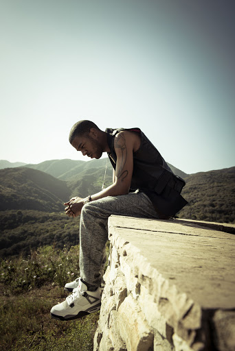 Young man sits on a stone wall overlooking a hilly landscape, wearing a black vest, grey sweatpants, and white sneakers on a sunny day.