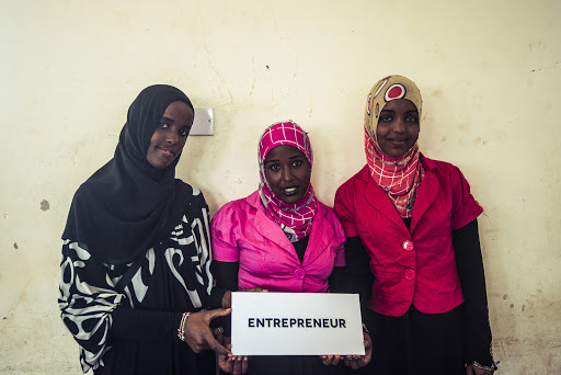Three women pose indoors, holding an 'Entrepreneur' sign, wearing colorful hijabs and outfits, against a neutral background.