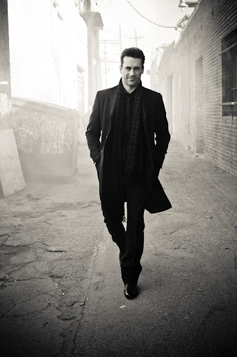 Monochrome shot of an adult man in a dark coat walking towards the camera on a city alleyway with buildings on both sides.