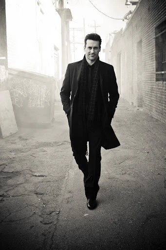 Monochrome shot of an adult man in a dark coat walking towards the camera on a city alleyway with buildings on both sides.
