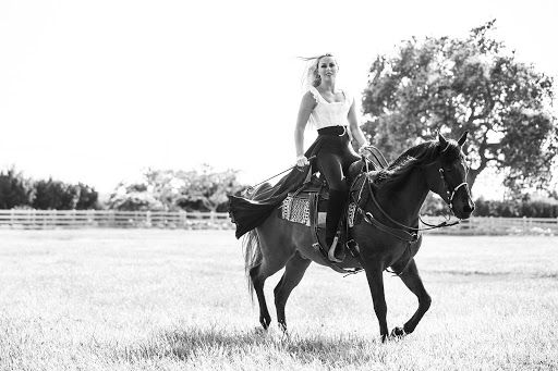 Monochrome photo of a woman riding a horse across a grassy field, wearing a fitted white top and dark riding pants, with trees in the background.