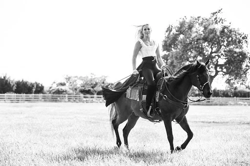 Monochrome photo of a woman riding a horse across a grassy field, wearing a fitted white top and dark riding pants, with trees in the background.