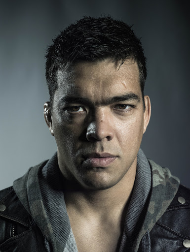 Dramatic portrait of a muscular adult male with short, dark hair wearing a leather jacket and hooded sweatshirt in a dimly lit indoor setting.