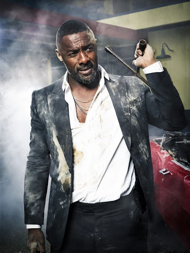 Man in a suit, covered in dirt, standing next to a wrecked red car, holding a lug wrench in a gritty, smoky garage setting.