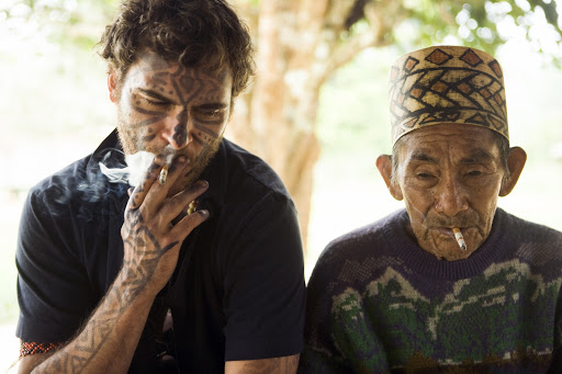 Two men, one with tribal face tattoos and the other elderly wearing a patterned hat, both smoking cigarettes in a blurry outdoor setting.