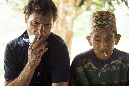 Two men, one with tribal face tattoos and the other elderly wearing a patterned hat, both smoking cigarettes in a blurry outdoor setting.