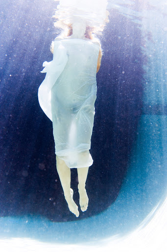 Underwater view of a woman in a long white dress with light shining through the water creating a serene and ethereal aesthetic.