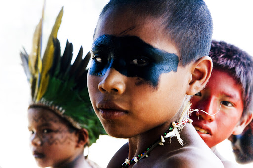 Close-up of an indigenous child with face paint and beaded necklace looking at the camera with other children in the background.