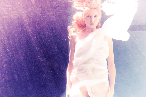 Underwater ethereal portrait of a fair-skinned adult woman draped in white fabric with flowing red hair, dreamlike and serene.