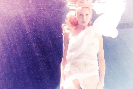 Underwater ethereal portrait of a fair-skinned adult woman draped in white fabric with flowing red hair, dreamlike and serene.