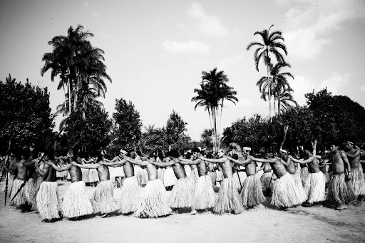 A line of men in grass skirts link arms and dance in a circle outdoors, surrounded by palm trees and foliage, in black and white.