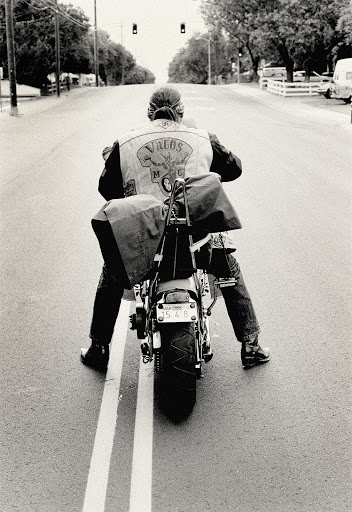 A biker wearing a leather jacket with saddlebags rides a motorcycle on an empty street in black and white on a sunny day.