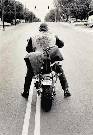 A biker wearing a leather jacket with saddlebags rides a motorcycle on an empty street in black and white on a sunny day.