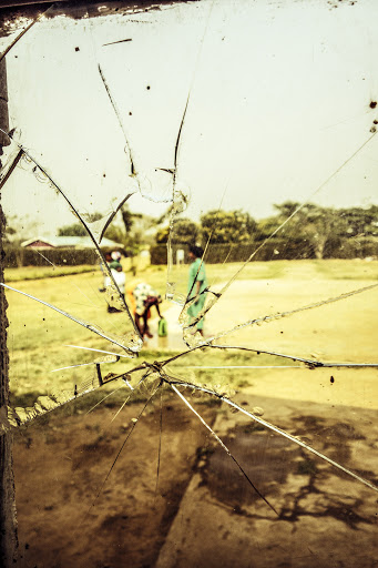 Cracked glass window with an outdoor view of three adults in casual clothing on a grassy lawn near trees and a residential structure.