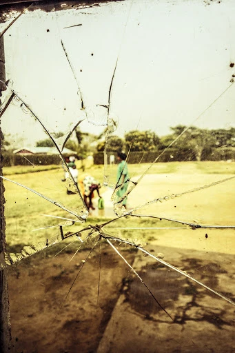 Cracked glass window with an outdoor view of three adults in casual clothing on a grassy lawn near trees and a residential structure.