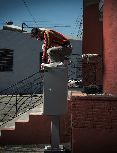 Young adult male crouches on a utility box in an urban alleyway wearing casual pants, colorful sweater, and white sneakers on sunny day.