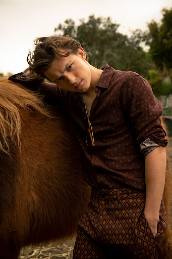 Portrait of a young adult man leaning against a brown horse in a rural outdoor setting, wearing a patterned shirt and pants.