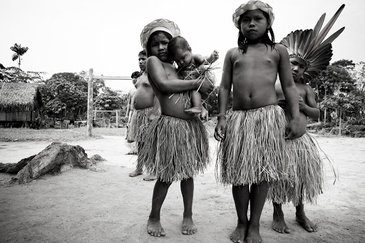 Black and white portrait of Indigenous children in grass skirts and headdresses. One child holds a baby. Huts and trees in background.