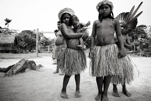 Black and white portrait of Indigenous children in grass skirts and headdresses. One child holds a baby. Huts and trees in background.