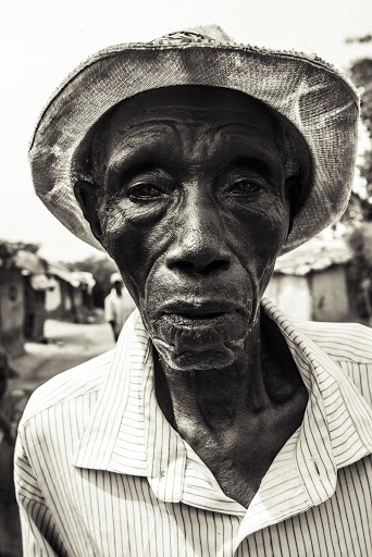 Monochrome portrait of an elderly African man wearing a hat and striped shirt, captured with a shallow depth of field in a rural setting.