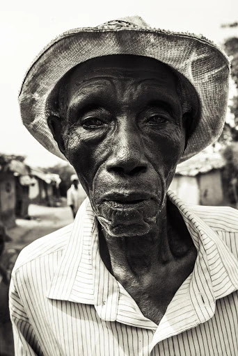 Monochrome portrait of an elderly African man wearing a hat and striped shirt, captured with a shallow depth of field in a rural setting.