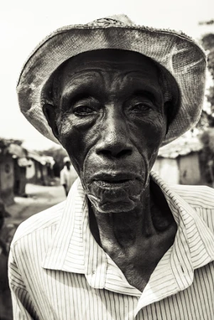 Monochrome portrait of an elderly African man wearing a hat and striped shirt, captured with a shallow depth of field in a rural setting.