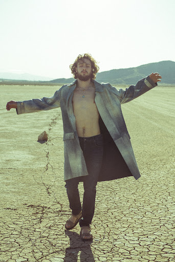 Adult male strikes a pose in an open field with mountains in the background. He wears a long coat and dark pants on cracked earth.