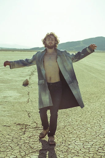 Adult male strikes a pose in an open field with mountains in the background. He wears a long coat and dark pants on cracked earth.