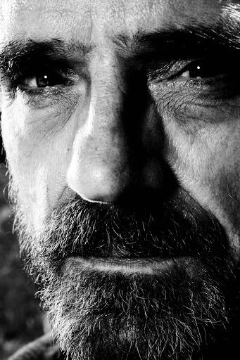 Close-up black and white portrait of an adult male with a beard, intense gaze, and visible skin texture, emphasizing depth and character.