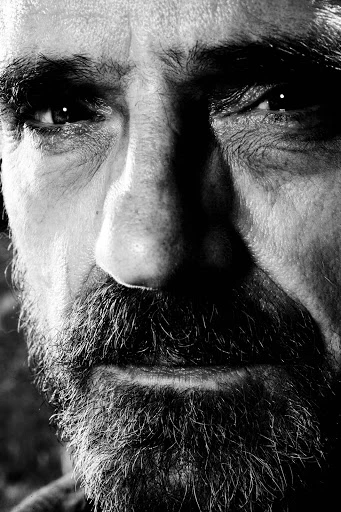 Close-up black and white portrait of an adult male with a beard, intense gaze, and visible skin texture, emphasizing depth and character.