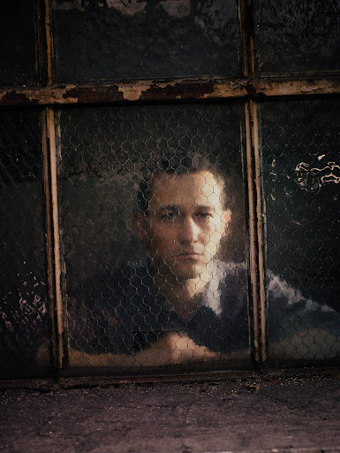 Indoor portrait of a man behind a wire mesh window. He leans an arm on the window ledge with a neutral expression on his face.