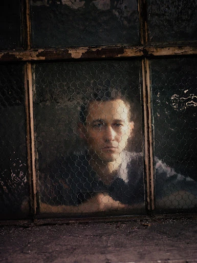 Indoor portrait of a man behind a wire mesh window. He leans an arm on the window ledge with a neutral expression on his face.