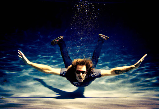 Man underwater in a swimming pool with arms outstretched wearing sunglasses and dark jeans photographed by Michael Muller