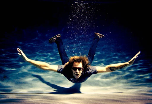 Man underwater in a swimming pool with arms outstretched wearing sunglasses and dark jeans photographed by Michael Muller