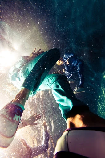 Low-angle shot of people swimming underwater in a bright, sunlit pool; one person wearing teal pants. Shot with eye-level angle of view.