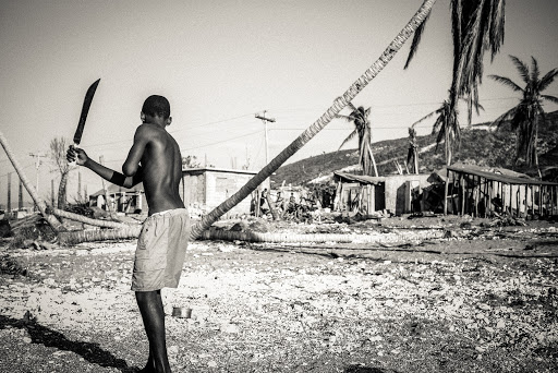 Monochromatic shot of a shirtless person holding a machete, standing in a debris-strewn area with damaged structures and palm trees.