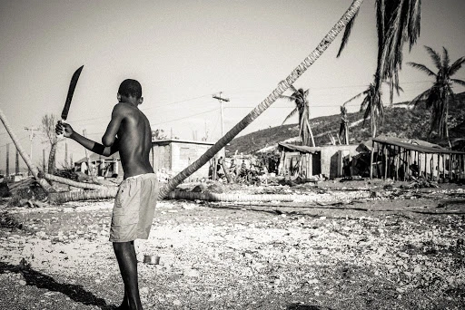 Monochromatic shot of a shirtless person holding a machete, standing in a debris-strewn area with damaged structures and palm trees.
