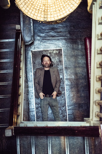 An adult male in a suit jacket and patterned pants stands in a stairwell, viewed from an overhead angle, with warm indoor lighting.