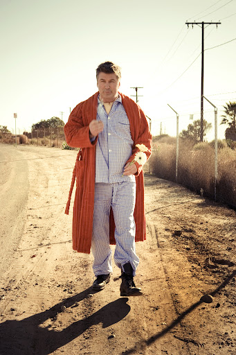 Adult male in robe and pajamas standing on a desert road with telephone poles and fence in background under bright sunlight.