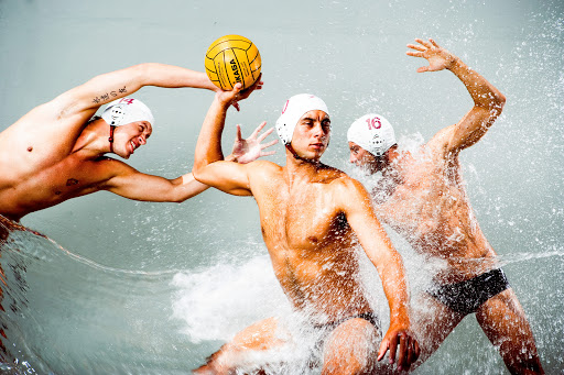 Three male water polo players compete for a yellow ball in a dynamic, splash-filled indoor scene, showcasing athleticism and competitive action.