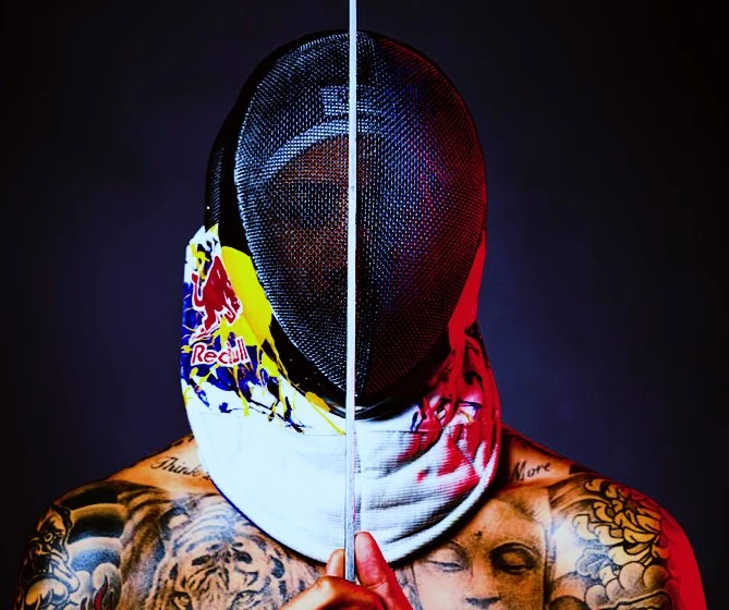 Close-up of a fencer wearing a Red Bull branded fencing mask and neck protector, partially obscuring their tattooed upper body.